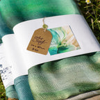Aqua coloured art scarves, folded with a price tag and product description on a grassy background
